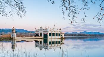 Un lago tranquilo con una casa flotante en el centro, rodeado de aguas apacibles y montañas al fondo.