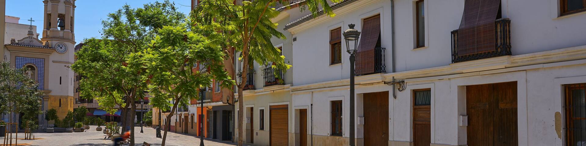 A sunny street with white buildings, a clock tower, and trees.