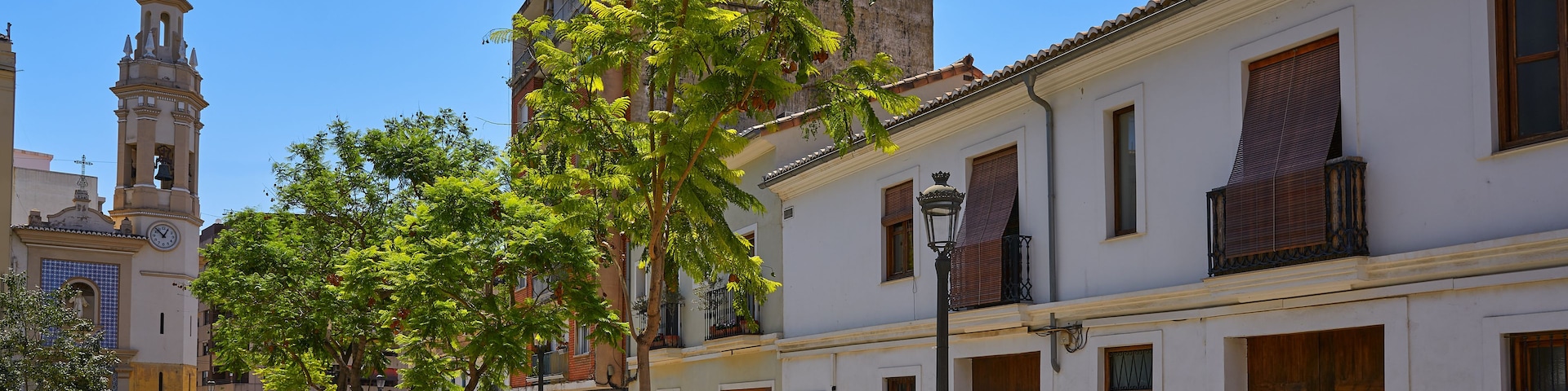 Rua ensolarada com edifícios brancos, torre do relógio e árvores.