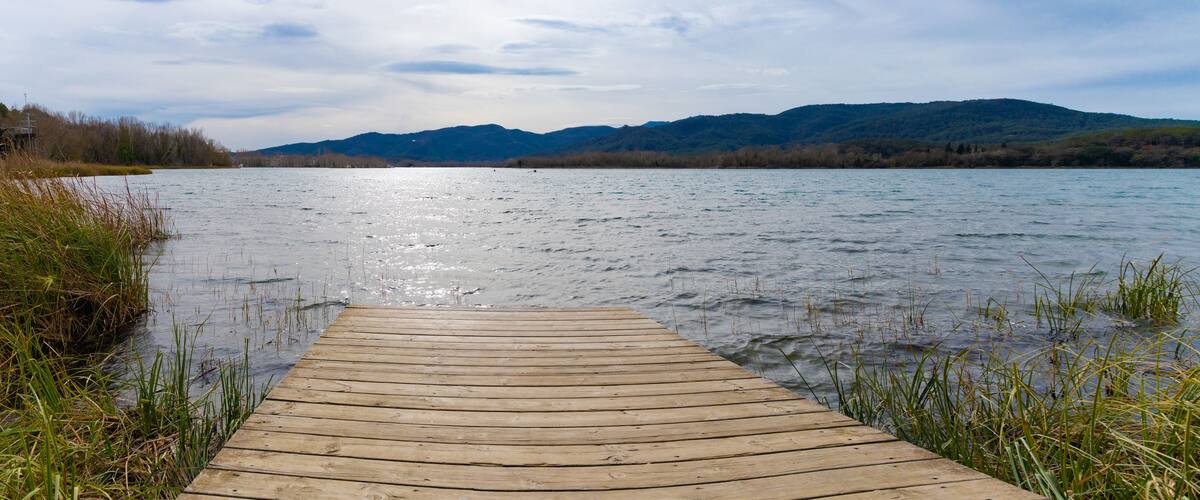 Un muelle de madera se extiende sobre las aguas del Lago Banyoles, ofreciendo una vista majestuosa de las colinas distantes bajo un cielo cubierto en el Pla de l'Estany, Cataluña, en invierno.