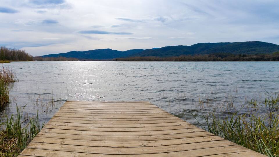 Un muelle de madera se extiende sobre las aguas del Lago Banyoles, ofreciendo una vista majestuosa de las colinas distantes bajo un cielo cubierto en el Pla de l'Estany, Cataluña, en invierno.