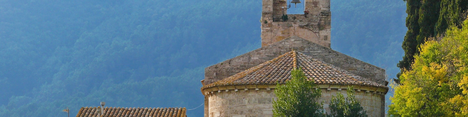 Una iglesia de piedra con un campanario, rodeada de exuberante vegetación y montañas al fondo.
