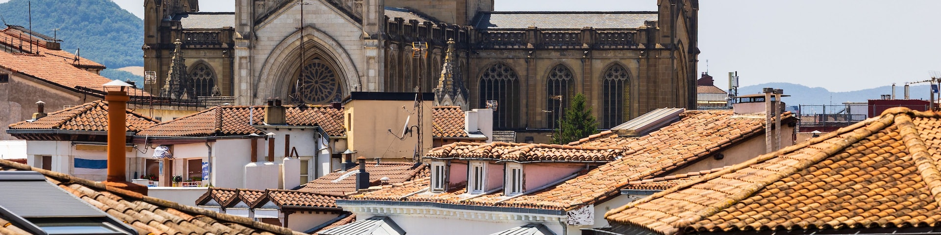A historic cathedral with Gothic architecture, surrounded by terracotta-tiled rooftops and a mountainous backdrop.