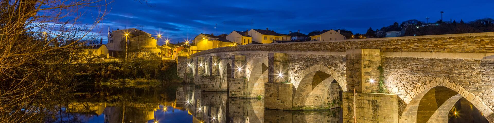 A stone bridge over a calm river at night, with buildings illuminated in the background.