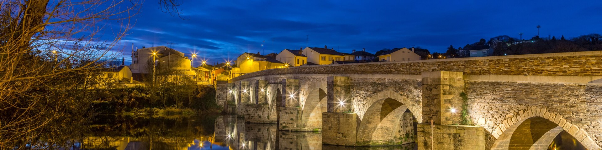 Uma ponte de pedra sobre um rio calmo à noite, com edifícios iluminados ao fundo.