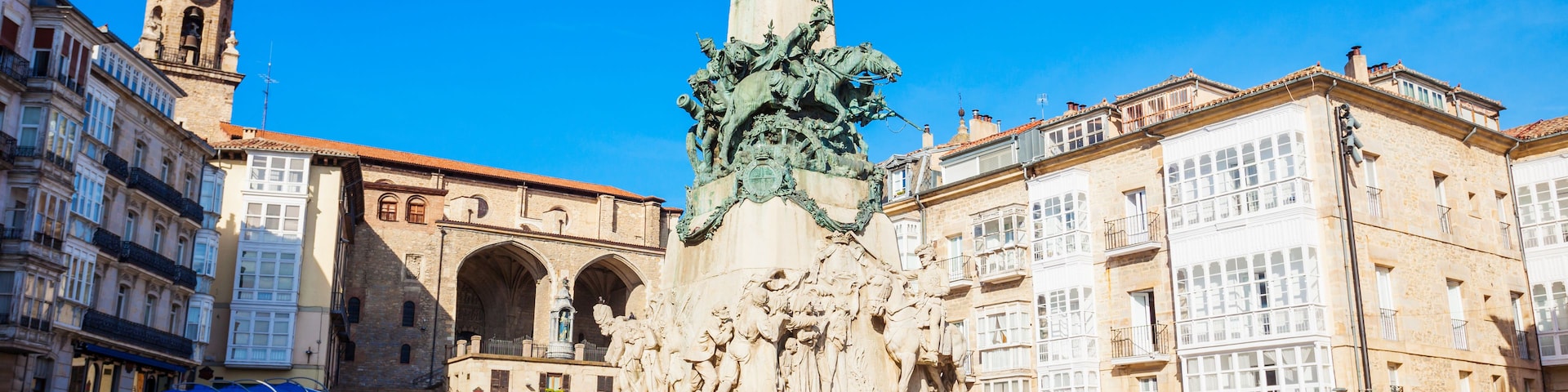 Uma praça com um monumento central, edifícios ao redor e uma torre do relógio.