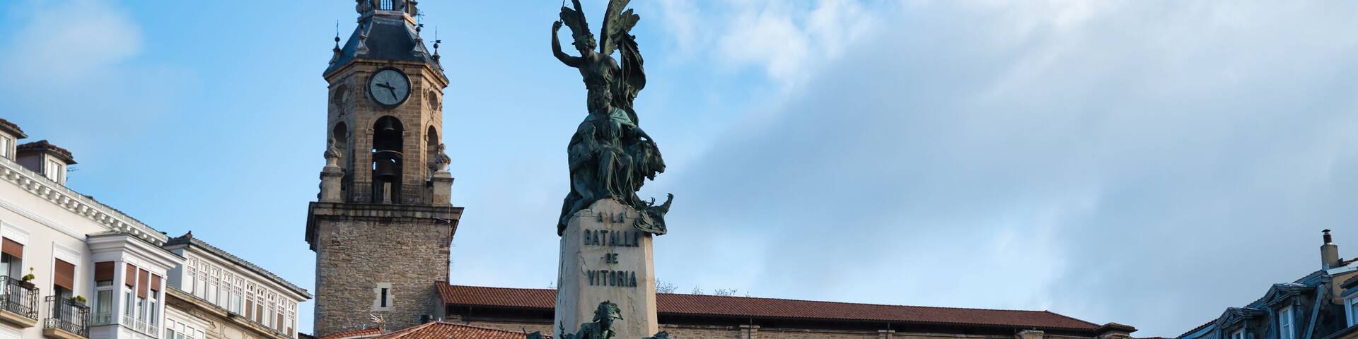 A historic square with a clock tower, a statue, and classical architecture.