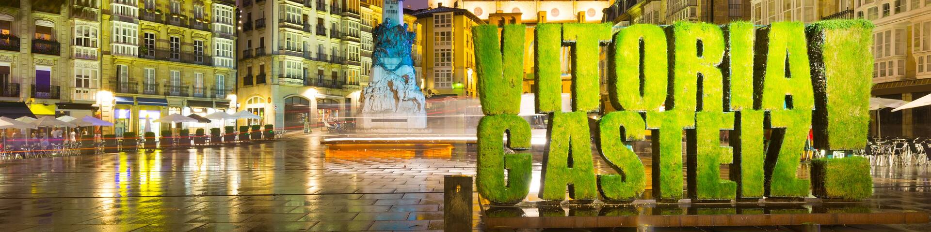 A nighttime city square with illuminated buildings, a statue, and the text 'VITORIA CASTELLO'.