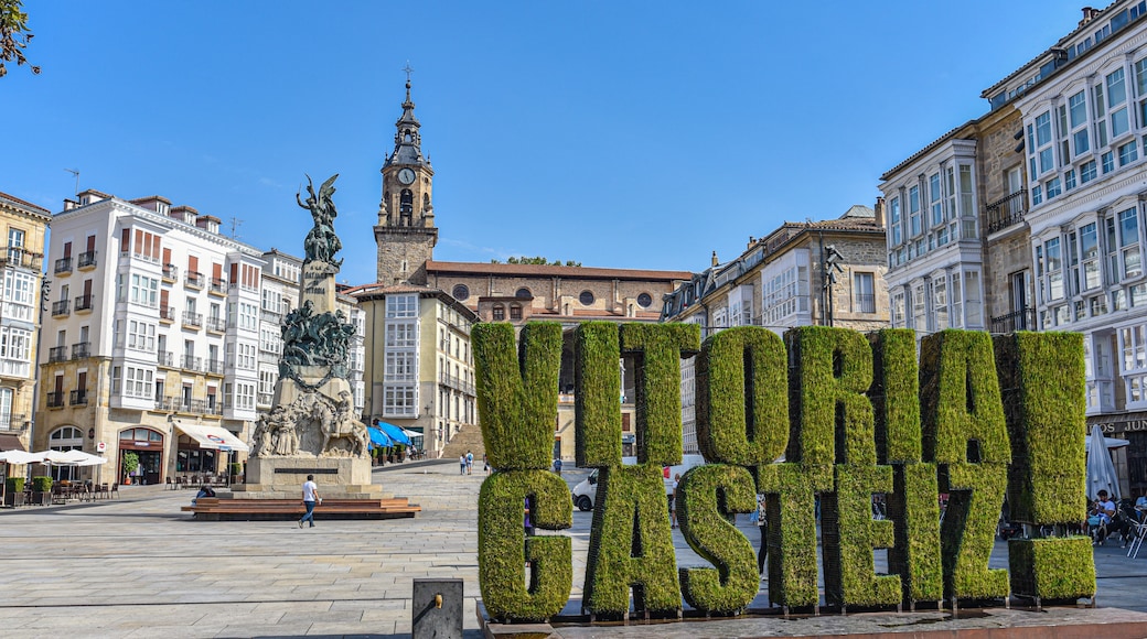 Une place agrémentée d’un grand panneau vert sur lequel on peut lire « VITORIA GASTEIZ », entourée de bâtiments historiques et d’une tour de l’horloge.