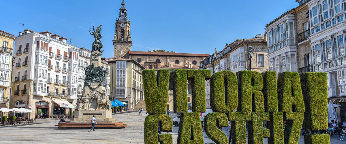 Una plaza con un gran cartel verde que dice 'VITORIA GASTEIZ', rodeada de edificios históricos y una torre del reloj.