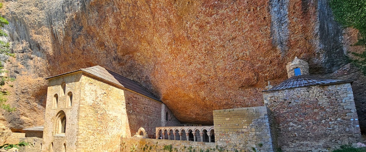 Un bâtiment historique en pierre, doté d’arches, avec une grande formation rocheuse en arrière-plan.