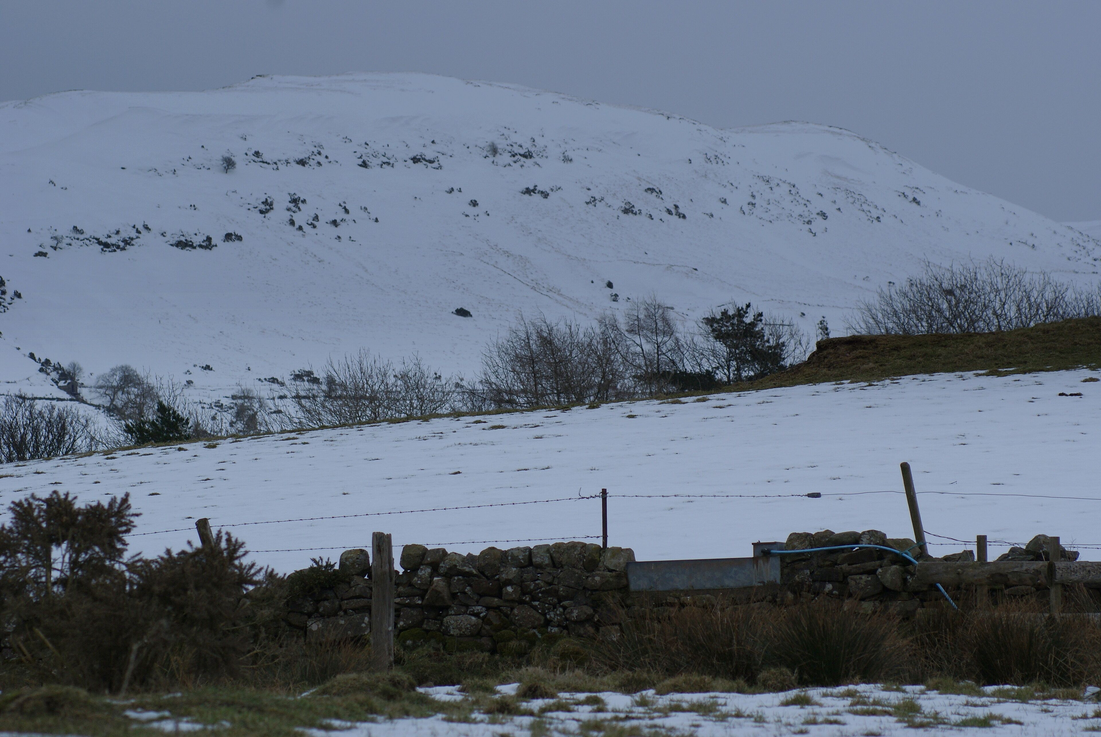 A snow-covered hill with a stone wall and barbed wire fence in the foreground.