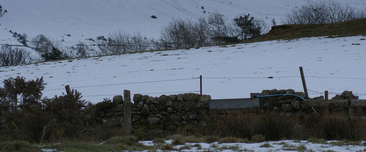 A snow-covered hill with a stone wall and barbed wire fence in the foreground.