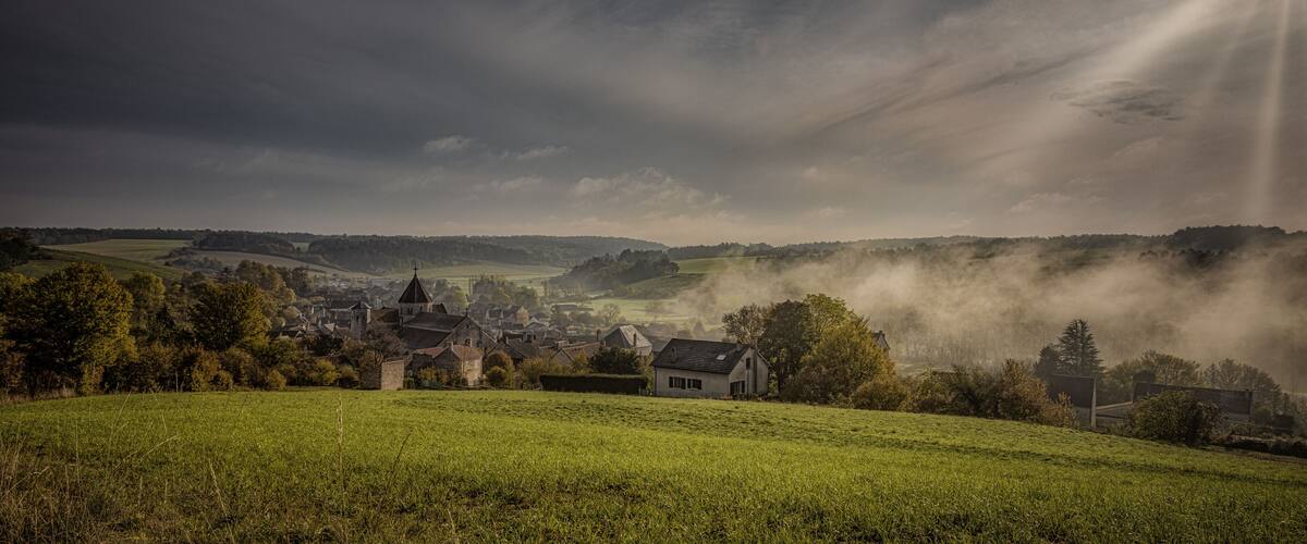 A village with houses and a church surrounded by green fields and hills under a cloudy sky.