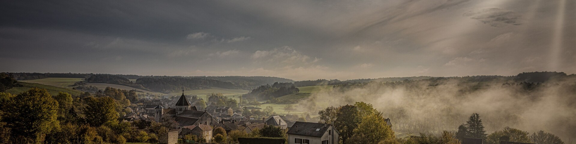 A village with houses and a church surrounded by green fields and hills under a cloudy sky.