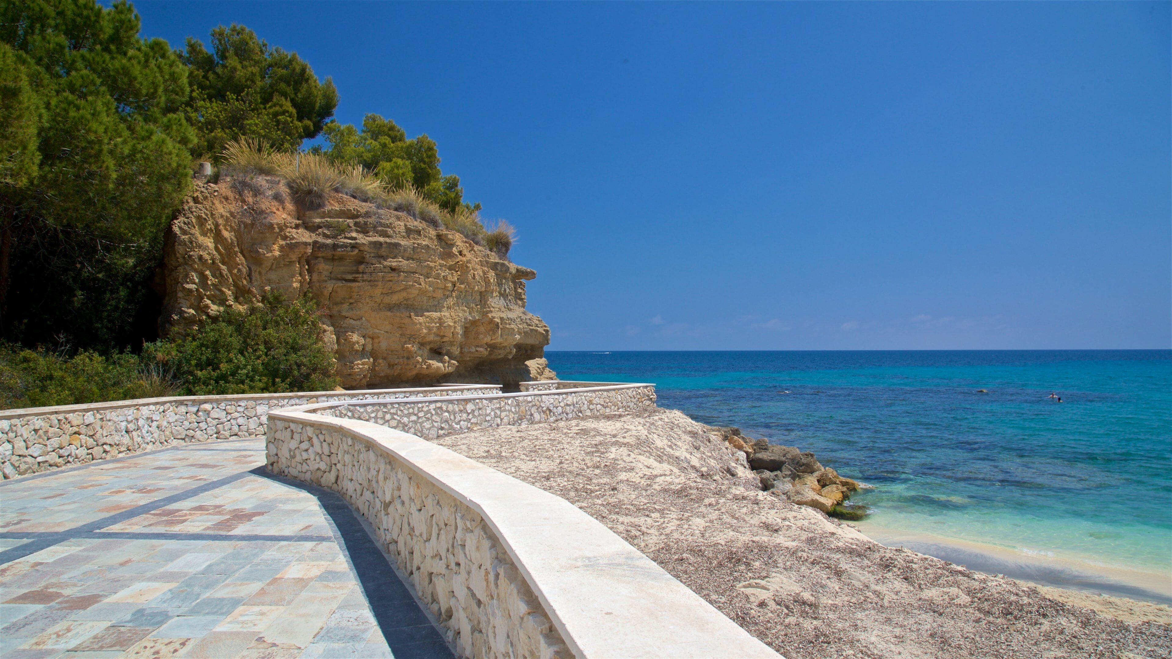 A coastal walkway with a stone wall, a rocky cliff, and the sea.