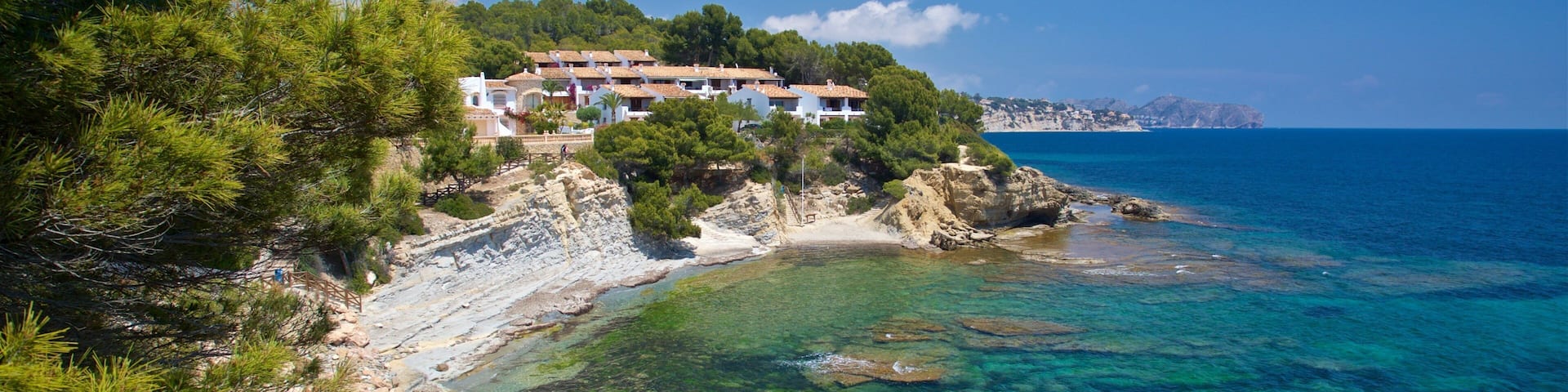 A coastal landscape with clear turquoise waters, rocky shoreline, pine trees, and a white building with red-tiled roofs on a hillside.