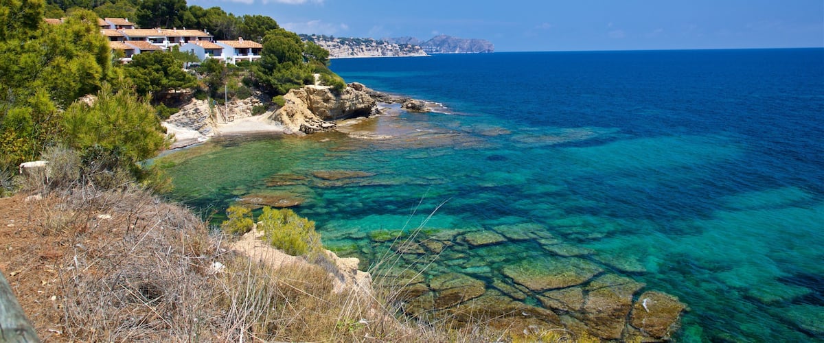 A coastal landscape with clear turquoise waters, rocky shorelines, and a cluster of houses on a hillside.