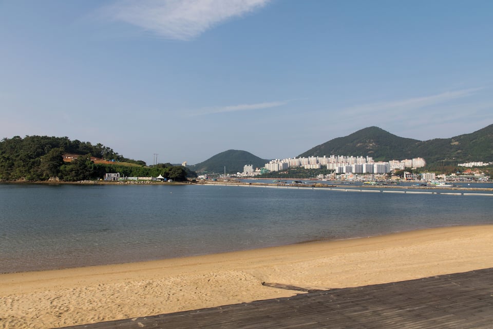 A beach with a wooden boardwalk, a calm sea, and a cityscape with buildings and hills in the background.