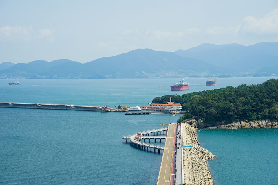A coastal city with a long pier extending into the sea, a bridge connecting the mainland to an island, and two large cargo ships in the distance.