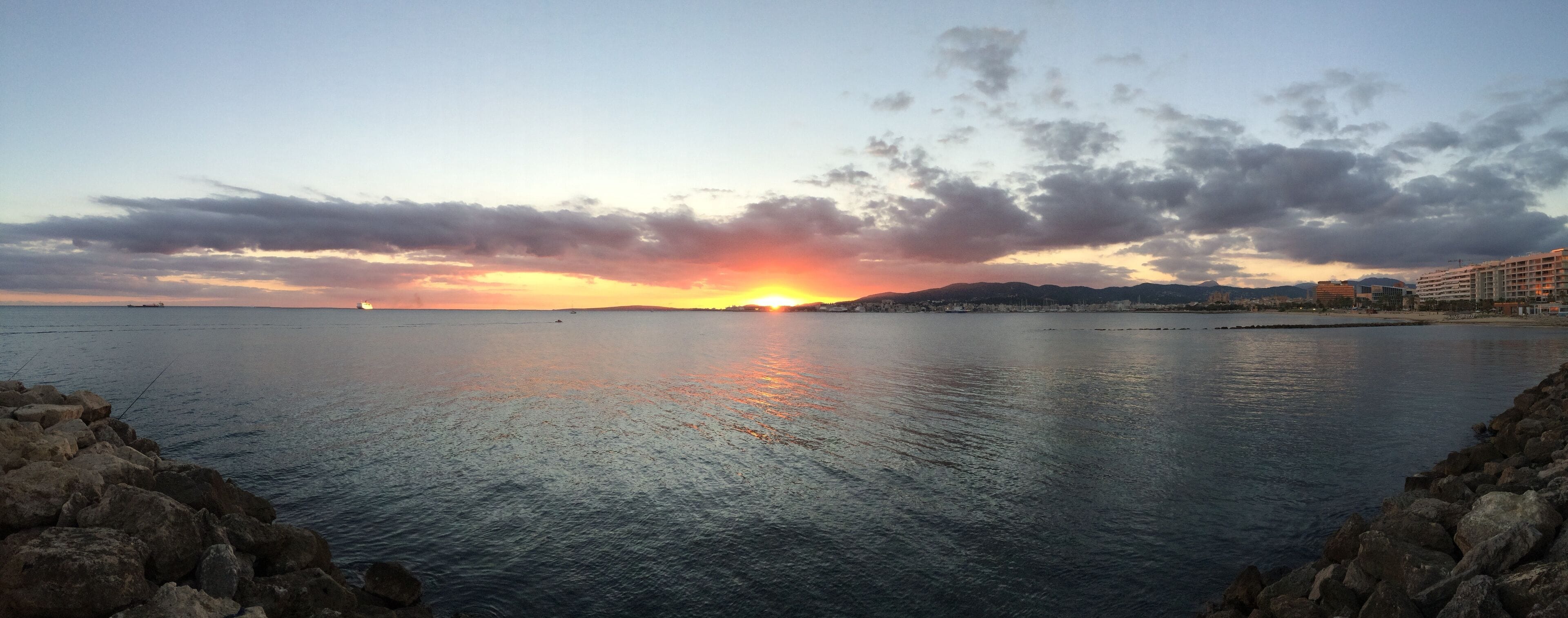A sunset over a calm sea with rocky shores and distant mountains.
