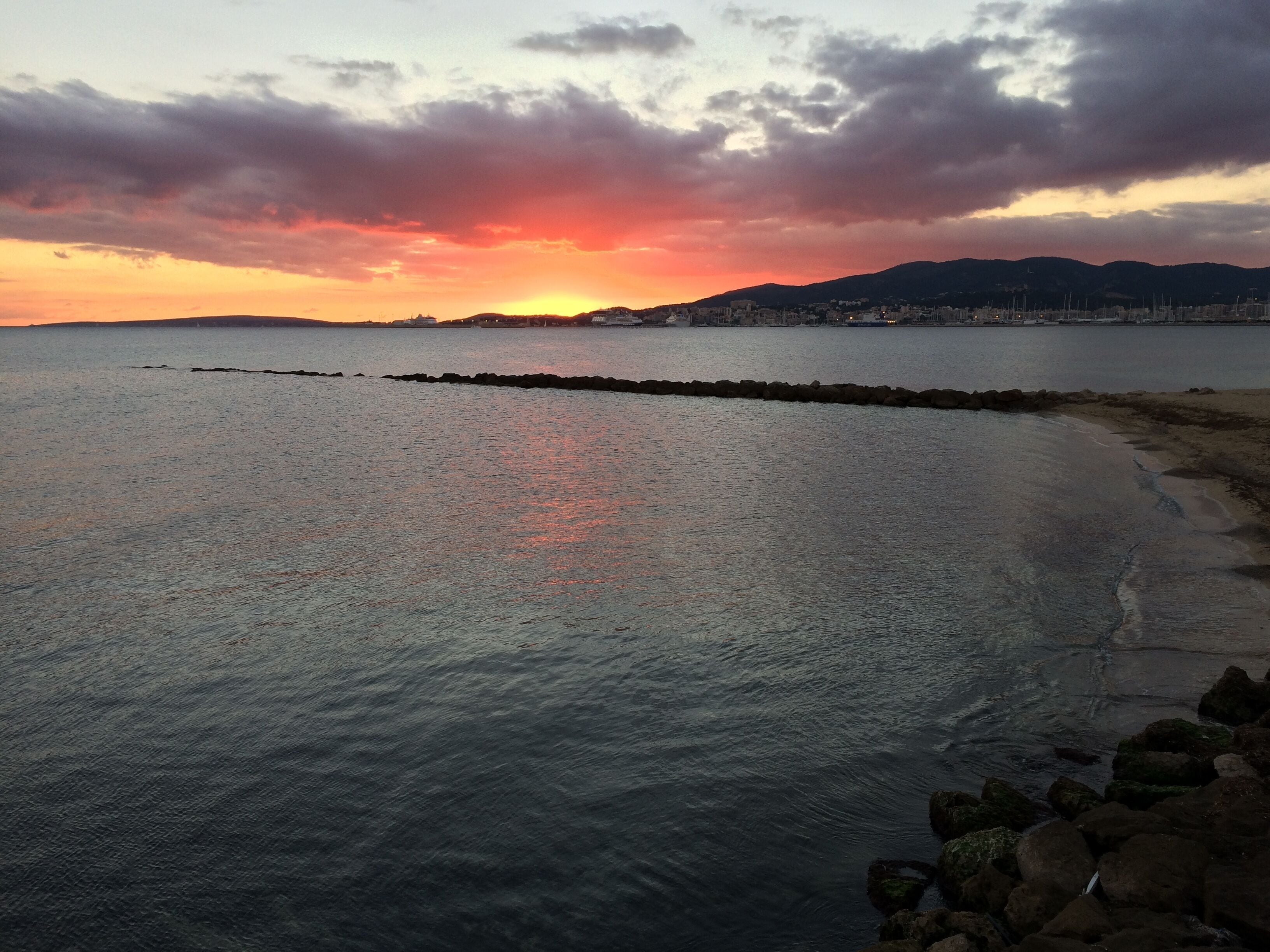 Um pôr do sol sobre um mar calmo, com uma costa rochosa e montanhas ao longe.