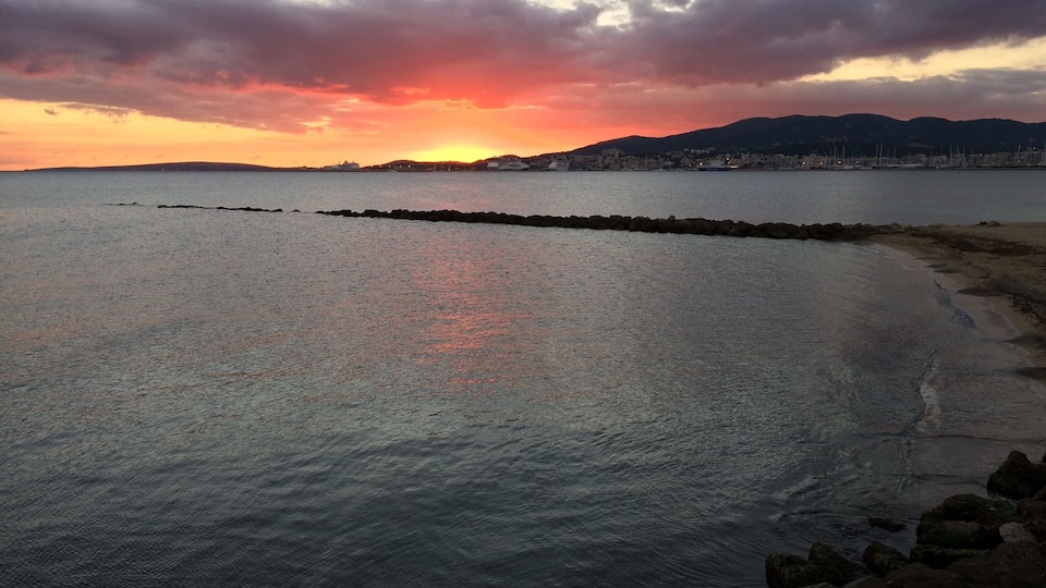 Um pôr do sol sobre um mar calmo, com uma costa rochosa e montanhas ao longe.
