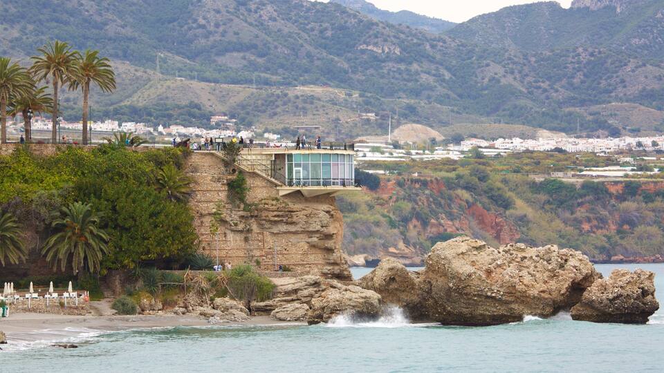 A cliffside building overlooking a coastal area with palm trees and a view of a town in the distance.