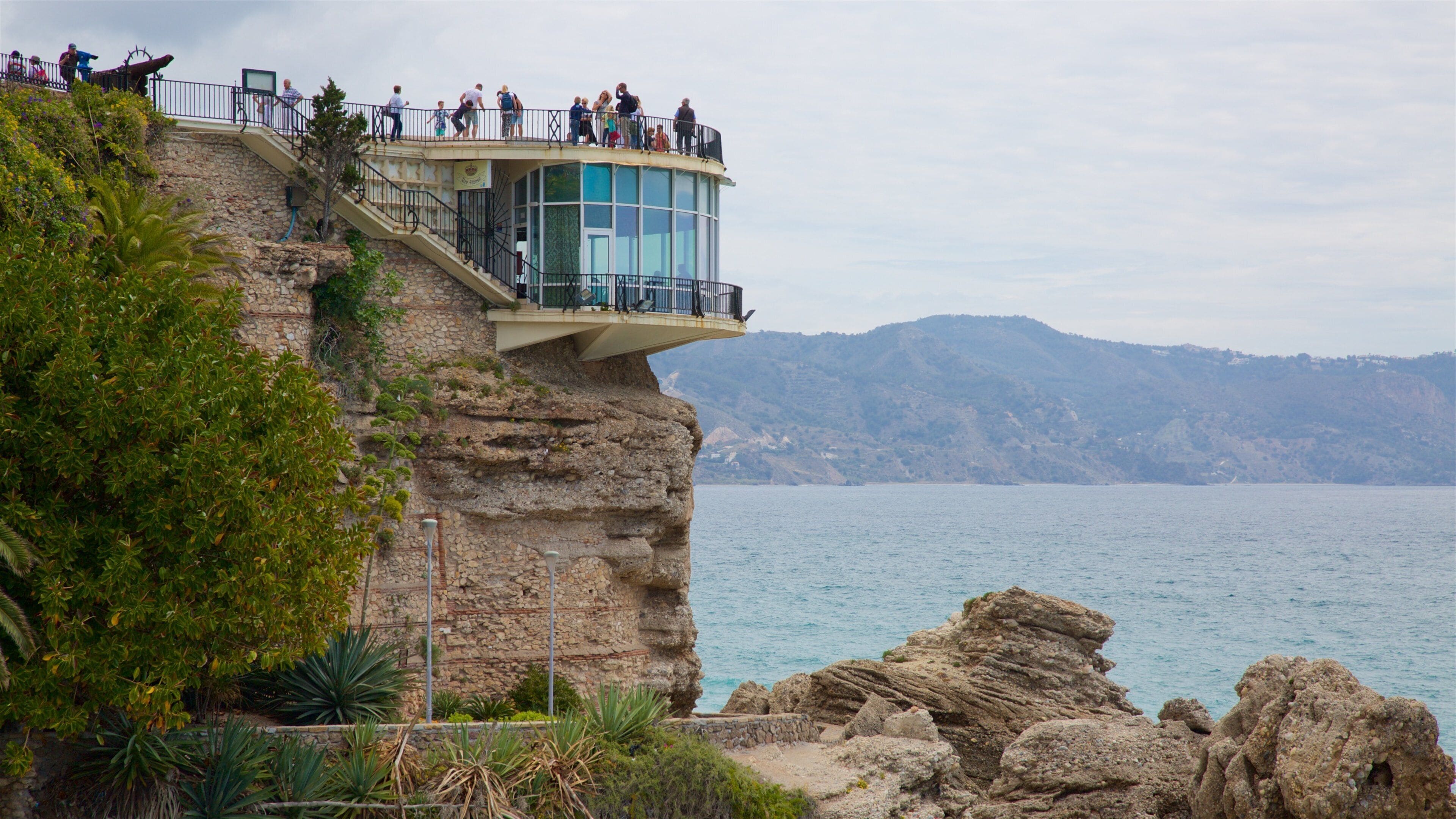A glass observation tower on a cliff overlooking a body of water with mountains in the background.
