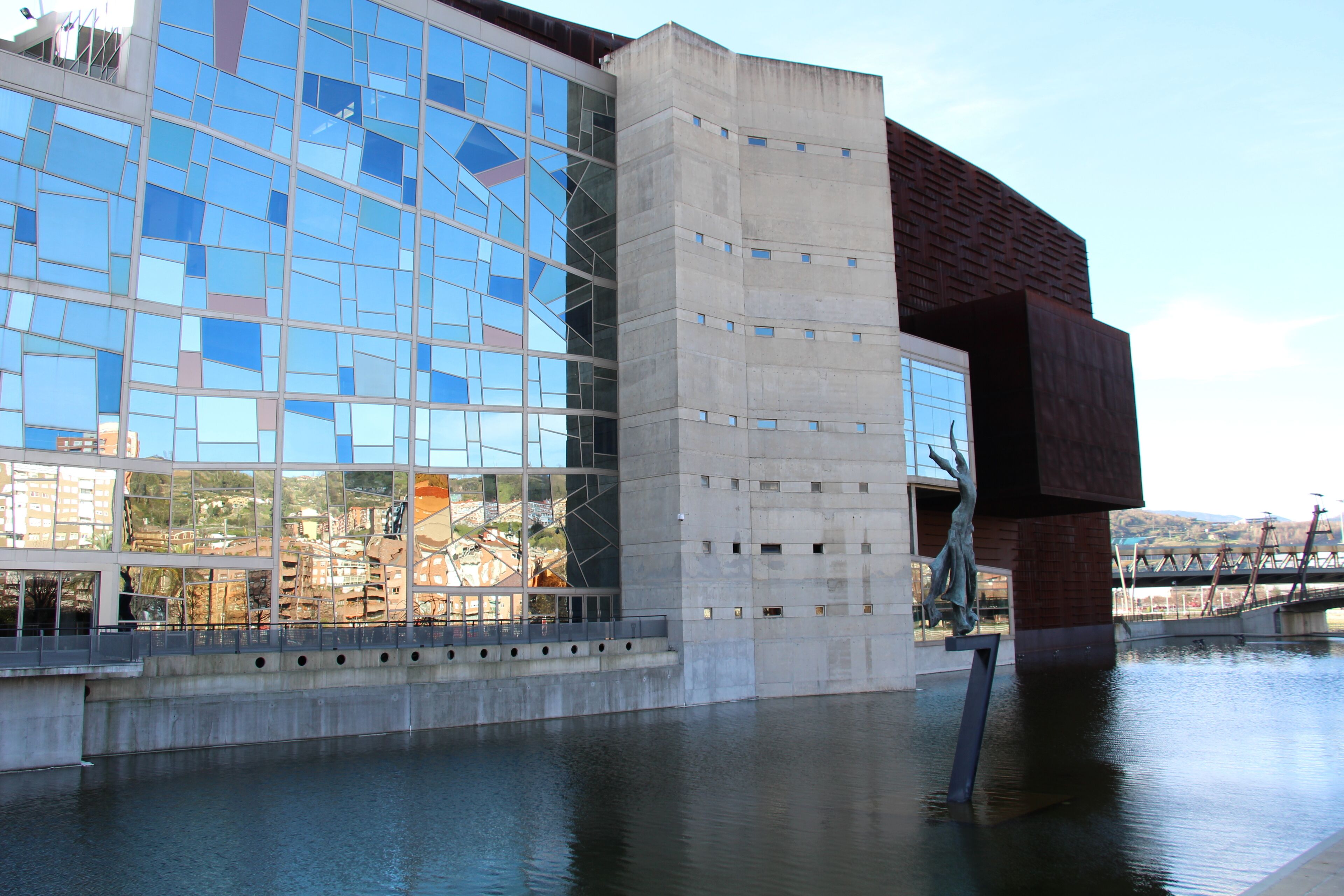 Un bâtiment moderne doté d’une façade en verre réfléchissant et d’une base en béton, situé au bord d’une étendue d’eau paisible.