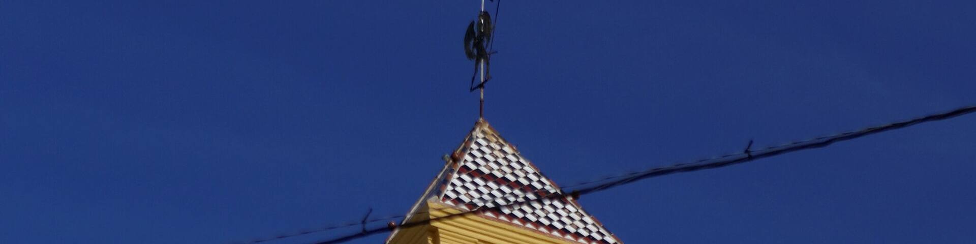 A yellow clock tower with a weather vane on top, set against a clear blue sky.