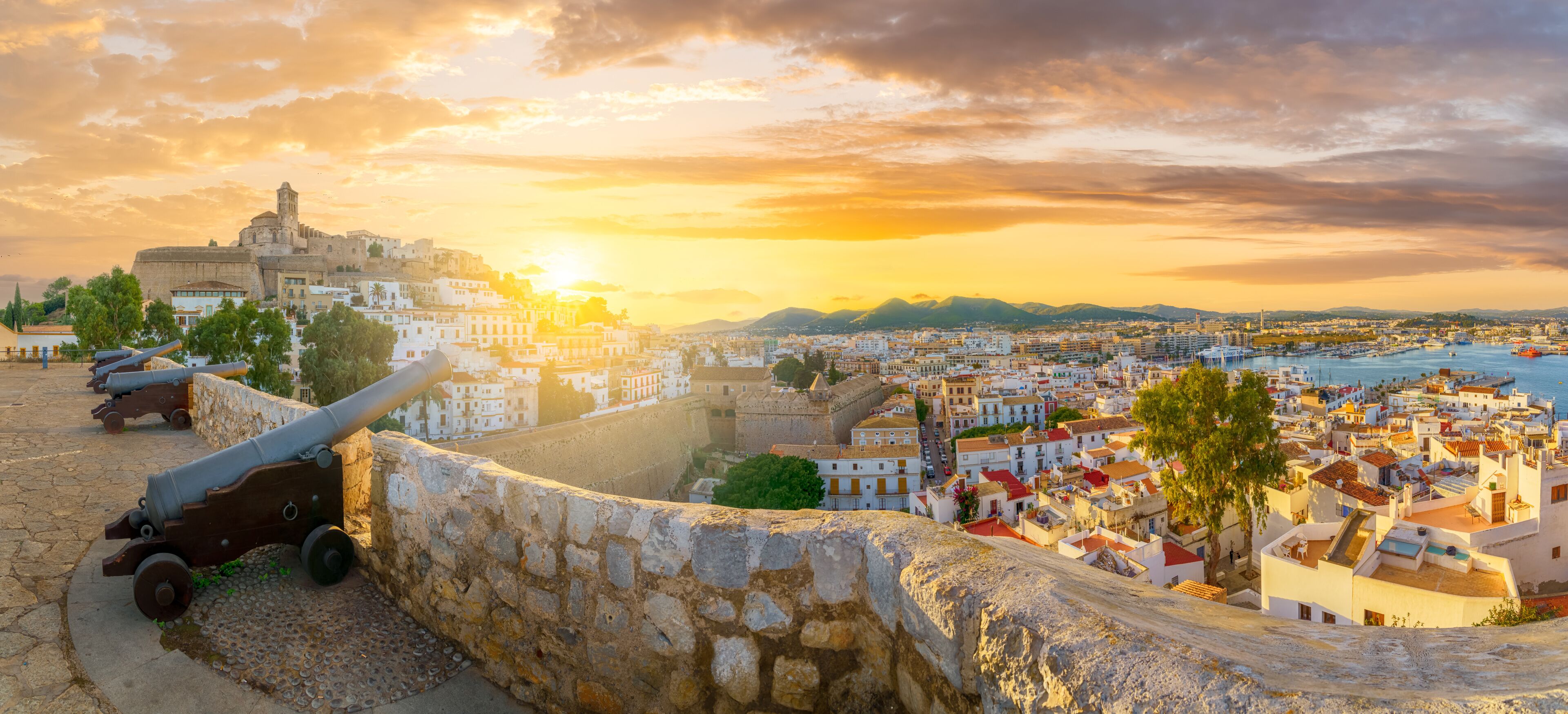 Un paysage urbain côtier avec des fortifications historiques, une église imposante et un coucher de soleil sur la mer.