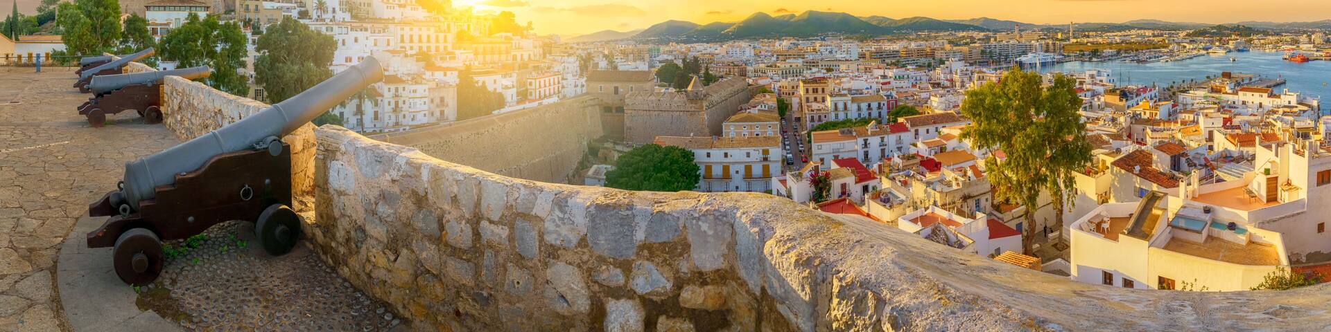 A coastal cityscape with historic fortifications, a prominent church, and a sunset over the sea.