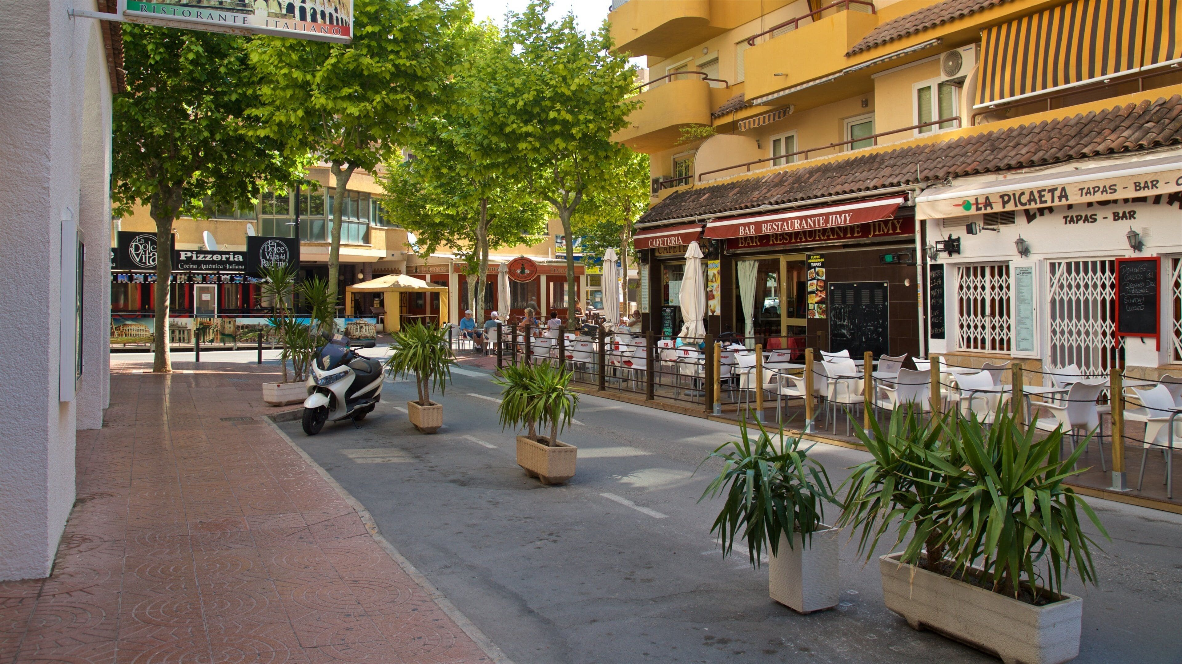 Eine Straße mit Außenbestuhlung verschiedener Cafés und Restaurants, ein Roller, der am Gehweg steht, und Pflanzkübel entlang des Bordsteins.