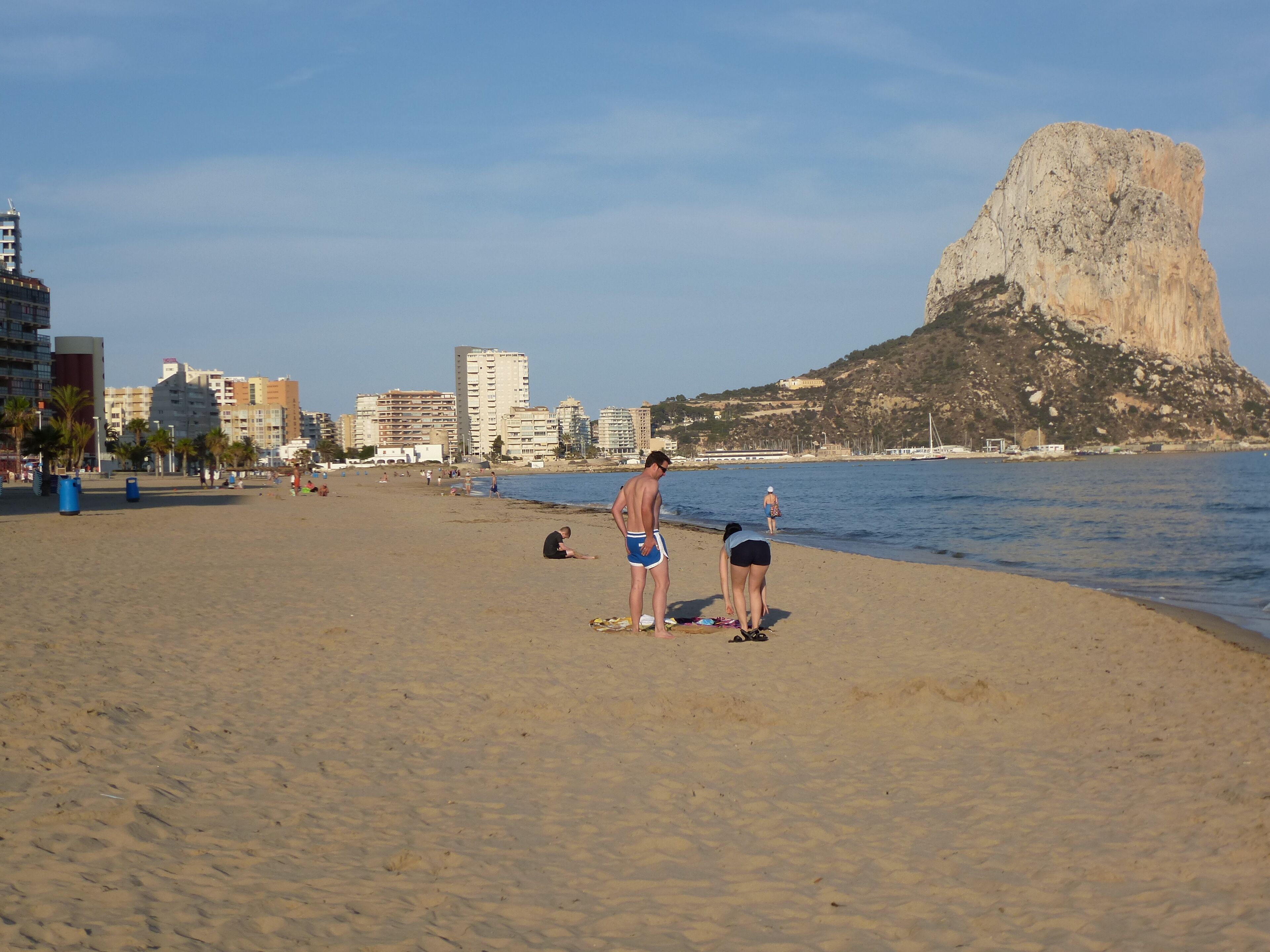Una playa con gente tomando el sol, una gran formación rocosa y edificios al fondo.