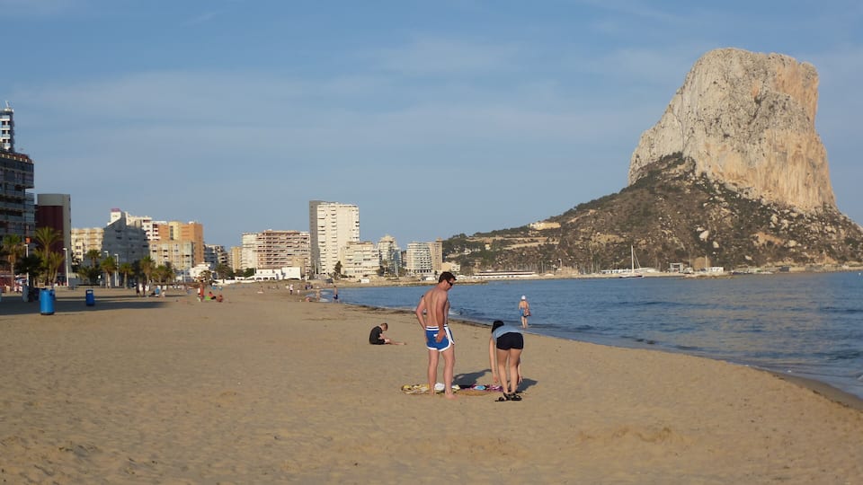 Una playa con gente tomando el sol, una gran formación rocosa y edificios al fondo.