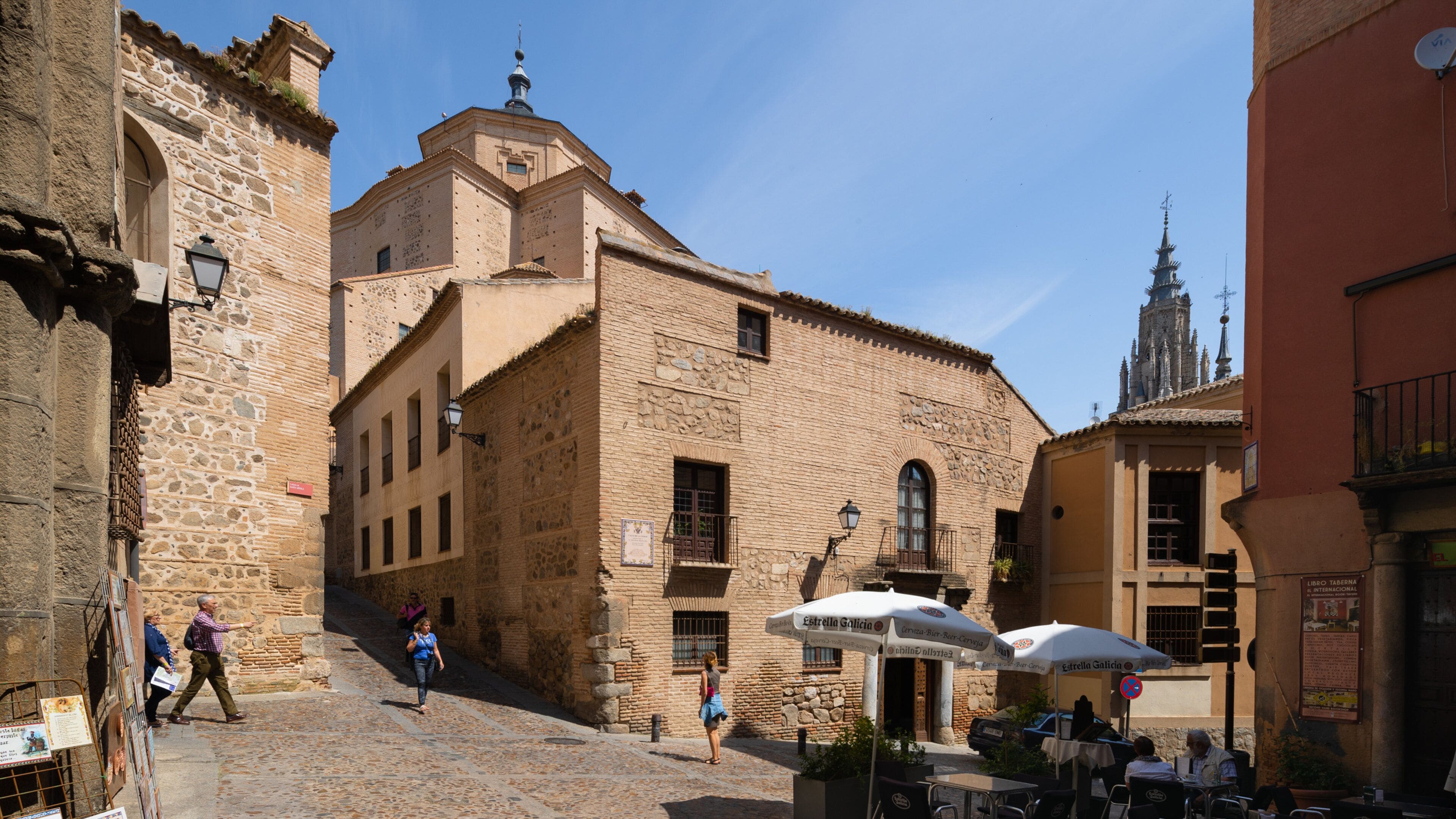 Un carrer pavimentat a una vila històrica amb edificis de pedra, una església i terrasses de café a l'aire lliure.