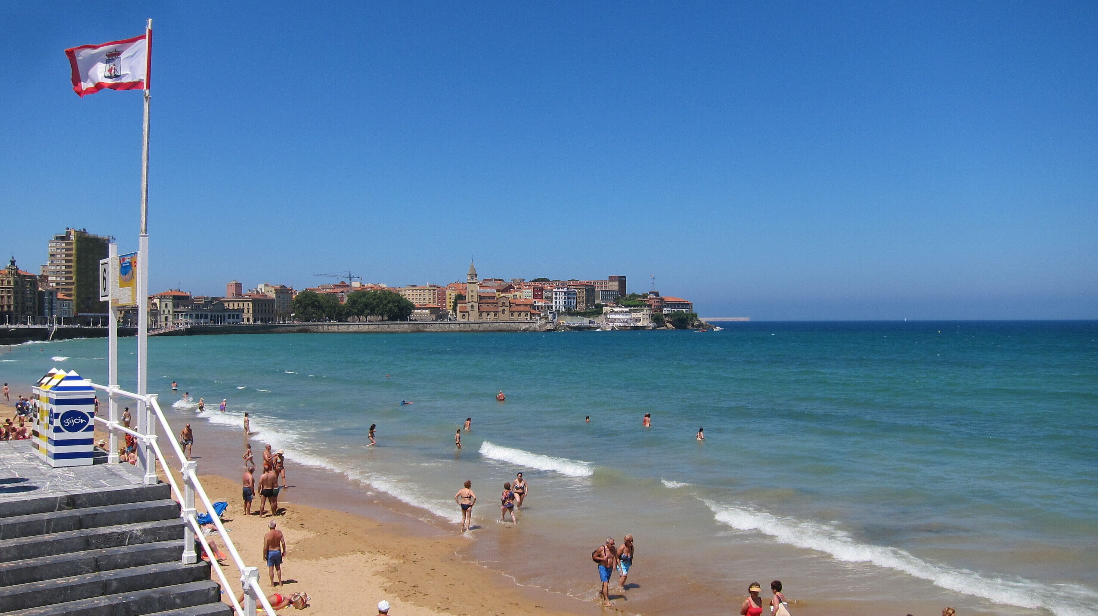 Una platja amb gent nedant i passejant al llarg de la riba, una bandera en un pal i edificis al fons.