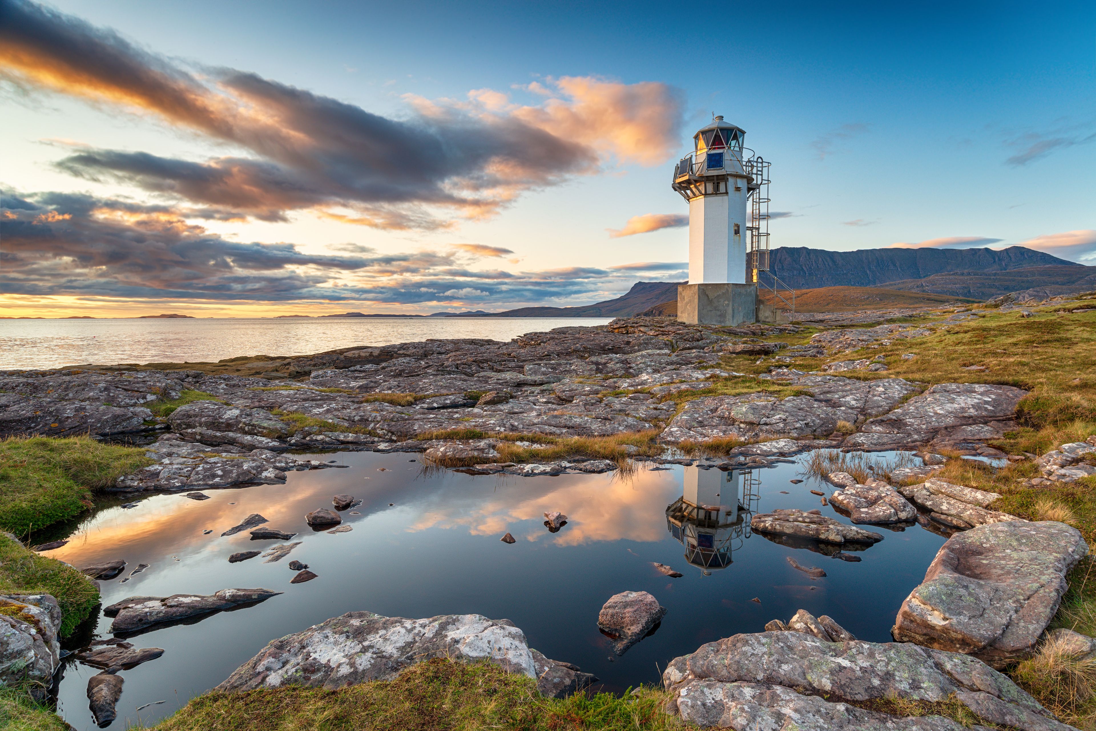 Un phare se dresse sur une côte rocheuse, et l’eau, calme et paisible, reflète le ciel.