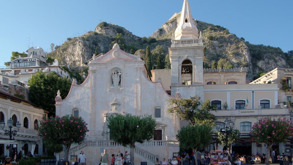 Une place historique, avec une église, un clocher et une chaîne de montagnes en arrière-plan.