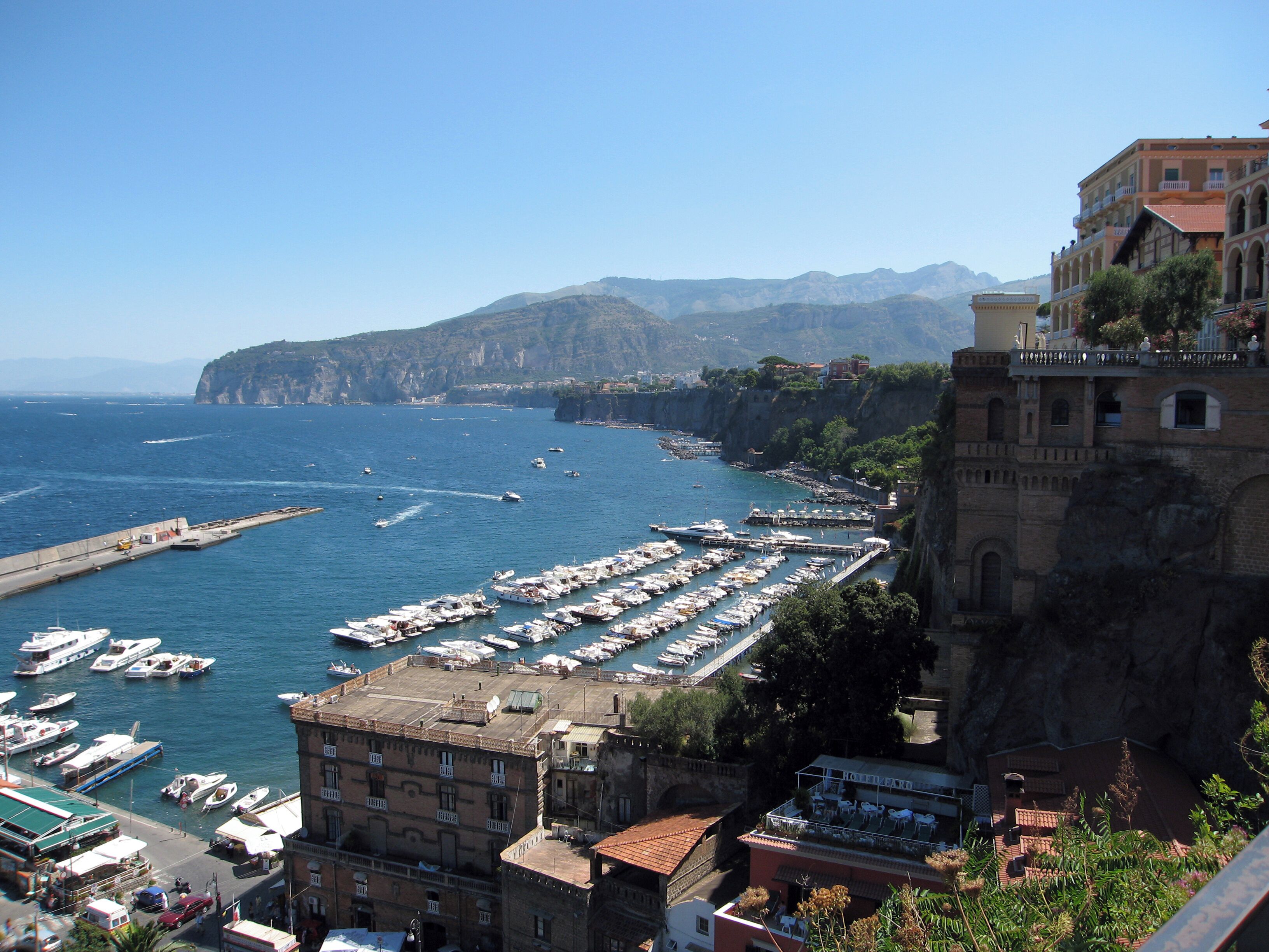 Una cittadina costiera con un porto turistico, edifici storici e scogliere sullo sfondo.