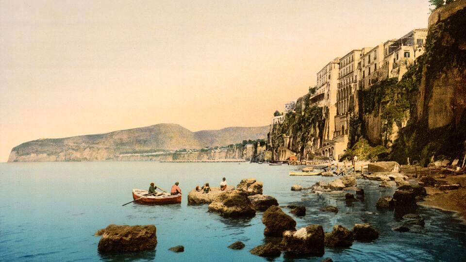 A coastal scene with rocky outcrops, calm sea, and buildings along the cliffside.