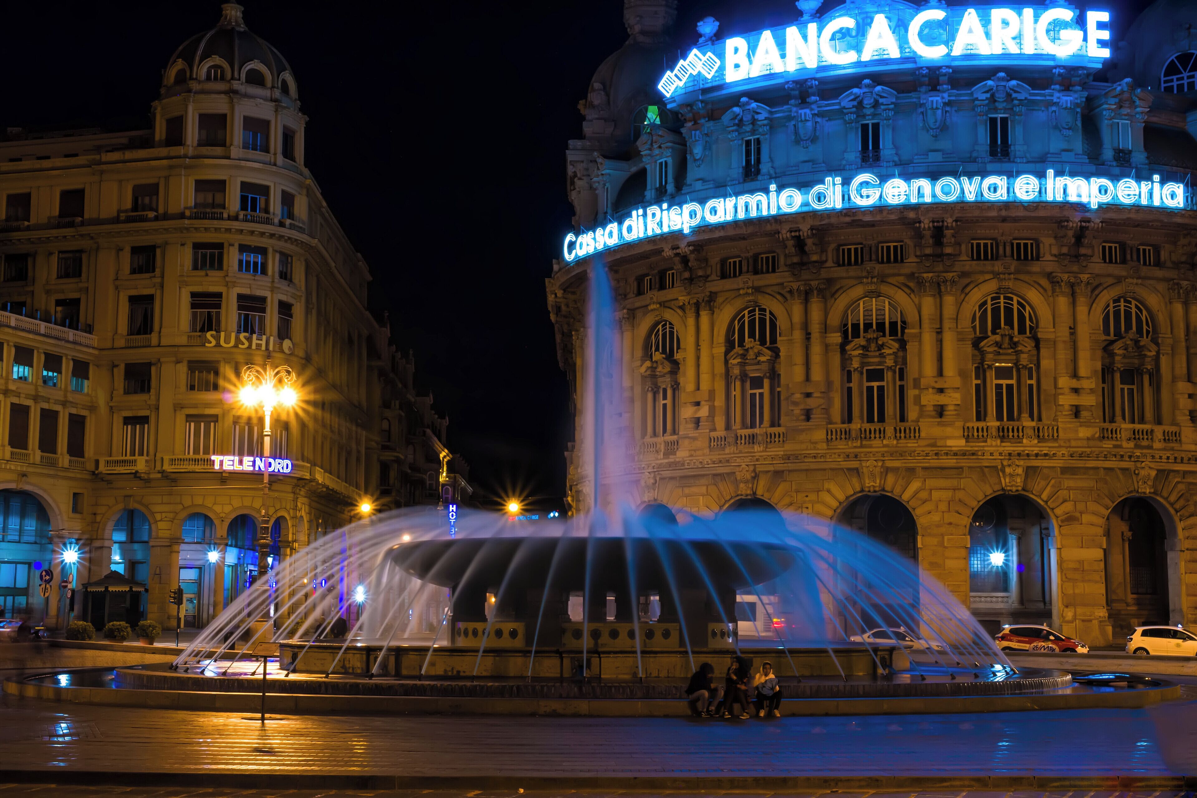 A historic building with illuminated signs, including "Banca Carige" and "Cassa di Risparmio di Genova e Imperia."