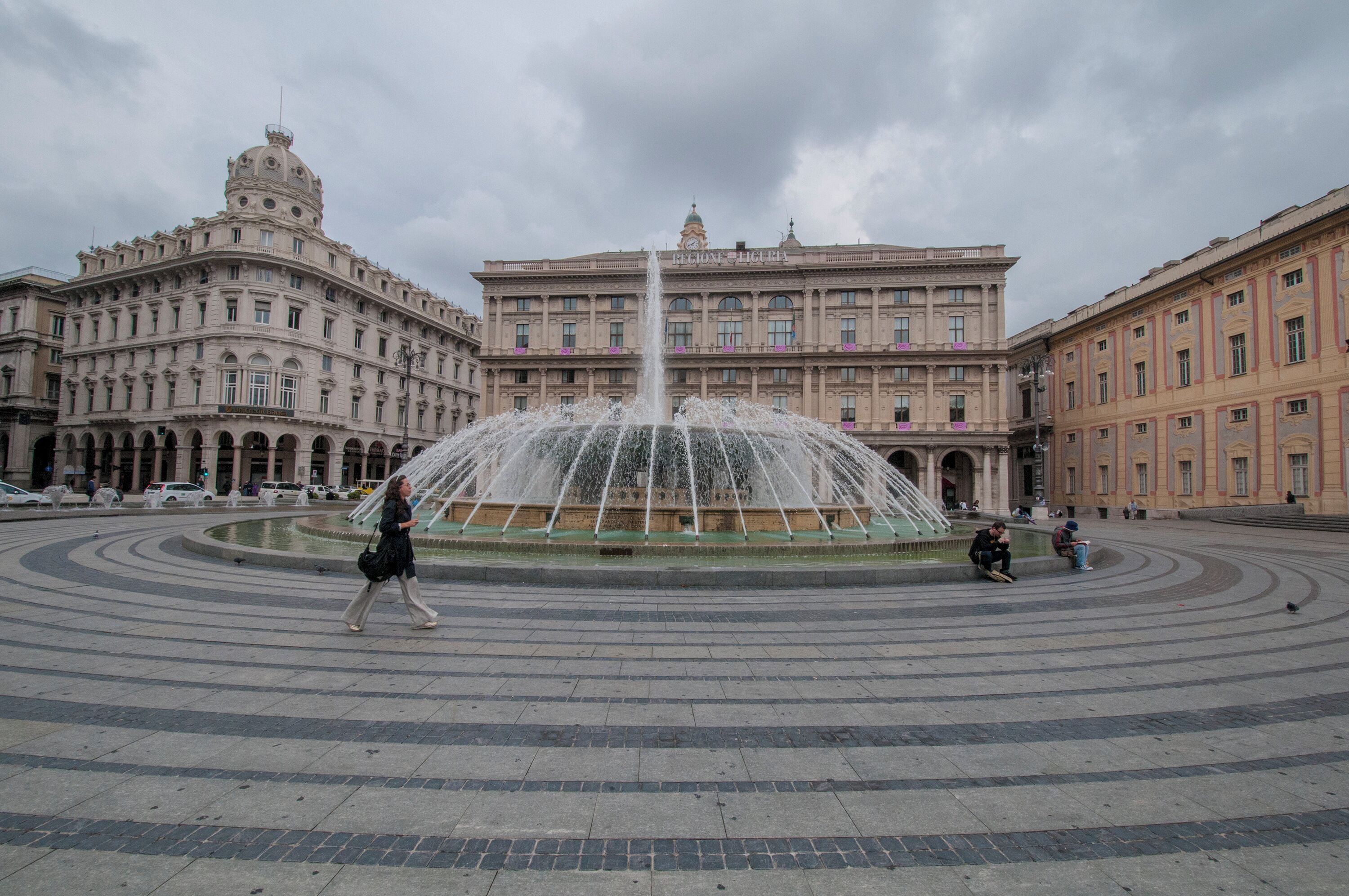 A large fountain in the center of a plaza with historic buildings on either side.