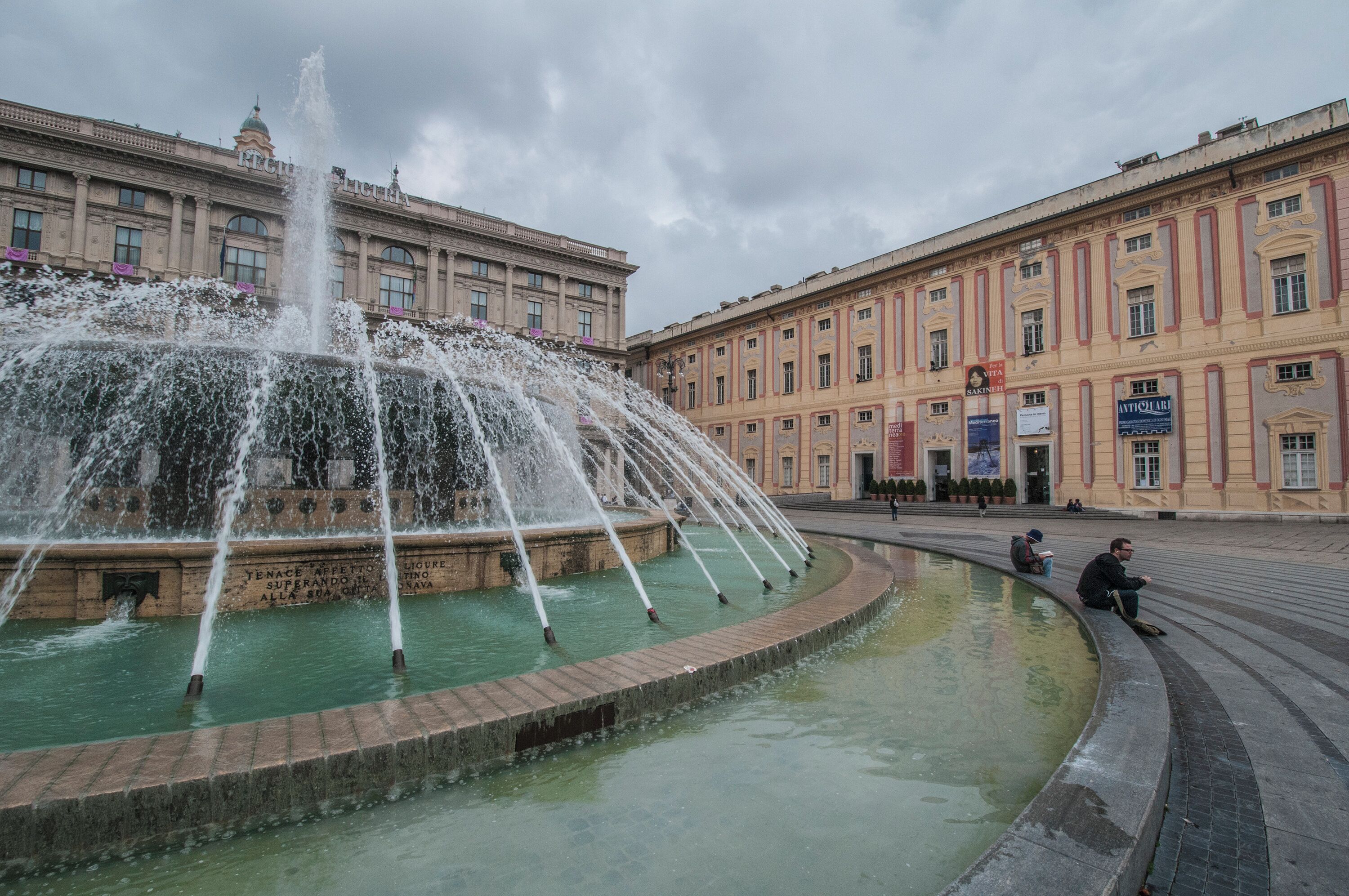 Una fontana storica con getti d'acqua multipli in una piazza pubblica, circondata da architettura classica.