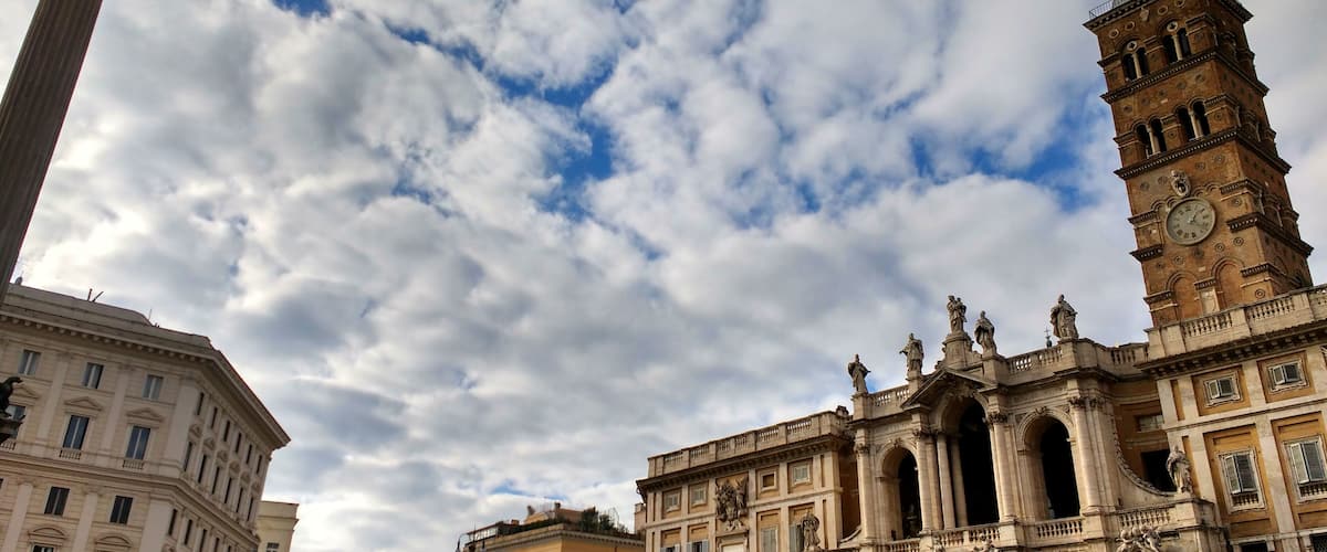 A historic square with a prominent clock tower, classical architecture, and a statue on a column.