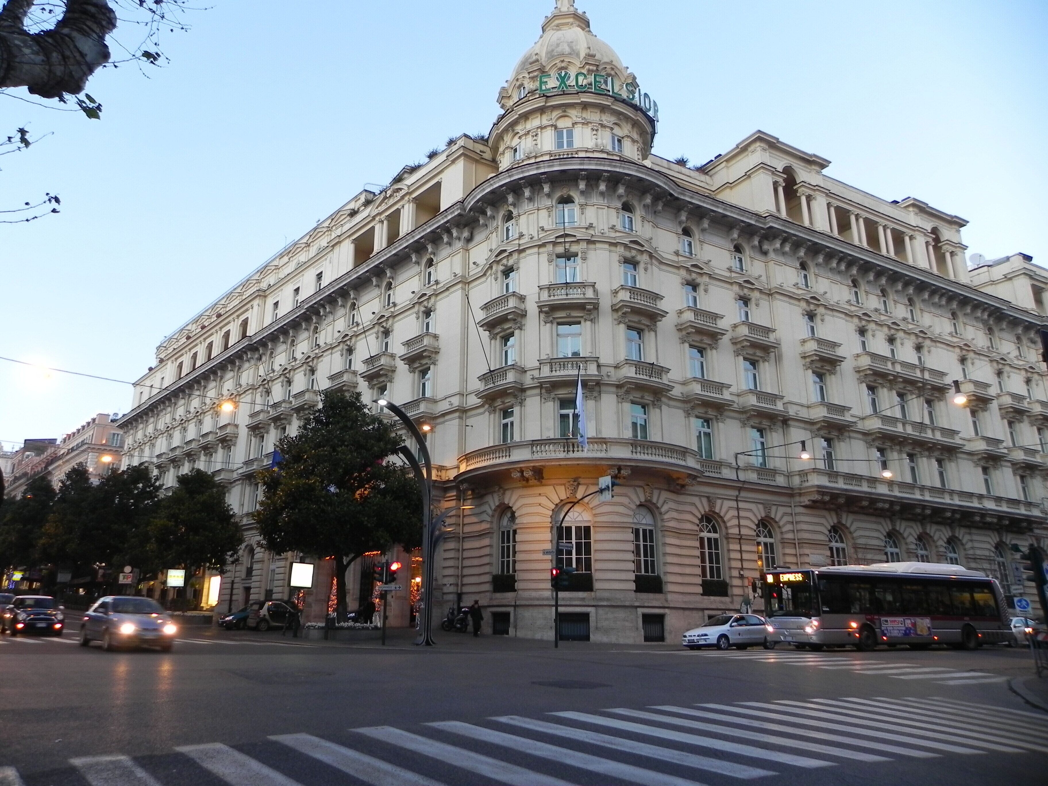 Um edifício grande e ornamentado, com cúpula e colunas, situado no cruzamento de uma cidade, com carros e um ônibus.