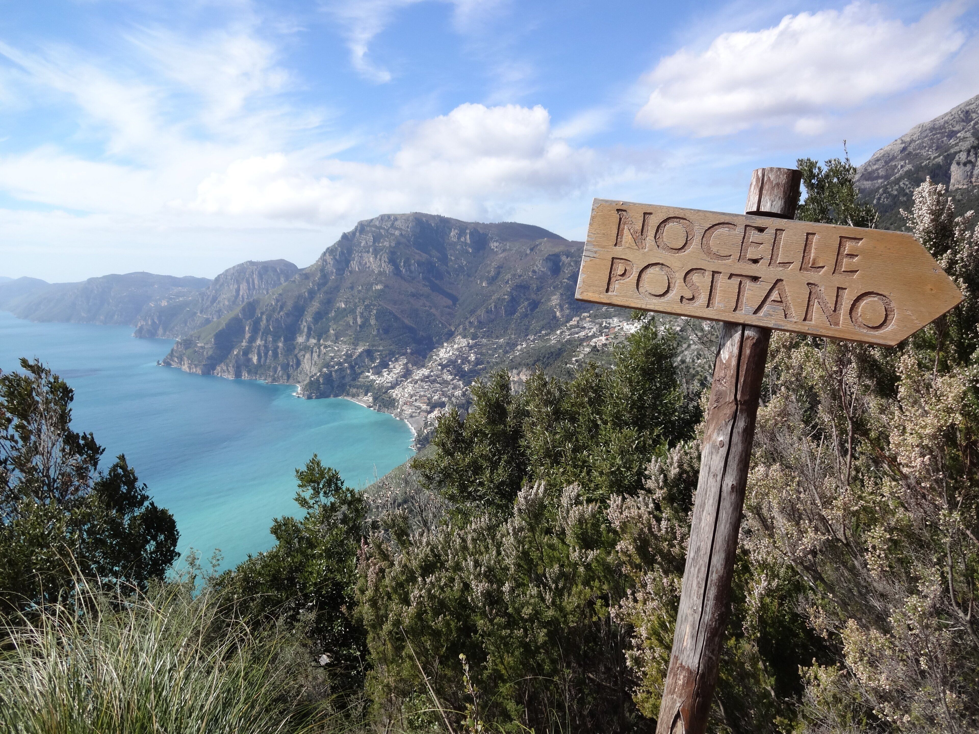 A wooden signpost with 'NOCCELLE POSTANO' carved on it, overlooking a coastal town with mountains in the background.