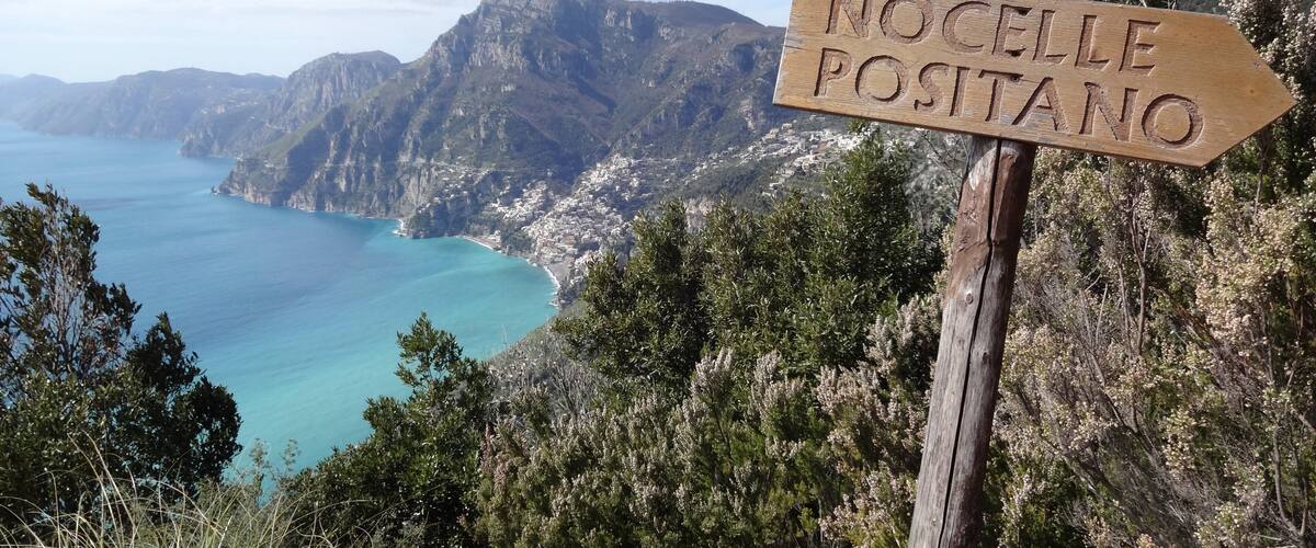 A wooden signpost with 'NOCCELLE POSTANO' carved on it, overlooking a coastal town with mountains in the background.