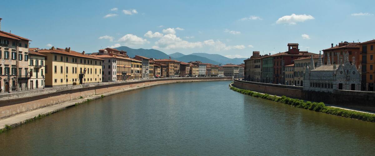 A river flanked by historic buildings and mountains in the background.